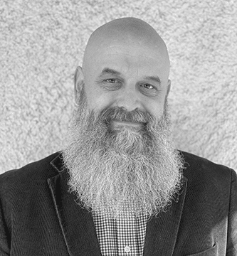 Smiling bald man with long gray beard wearing a checkered shirt and dark blazer against a textured background.