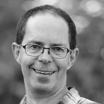 Smiling man with short hair and glasses wearing a plaid shirt with blurred background.