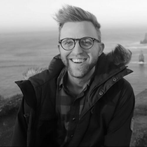 Smiling man with glasses and a jacket standing outdoors by the ocean with cliffs in the background.