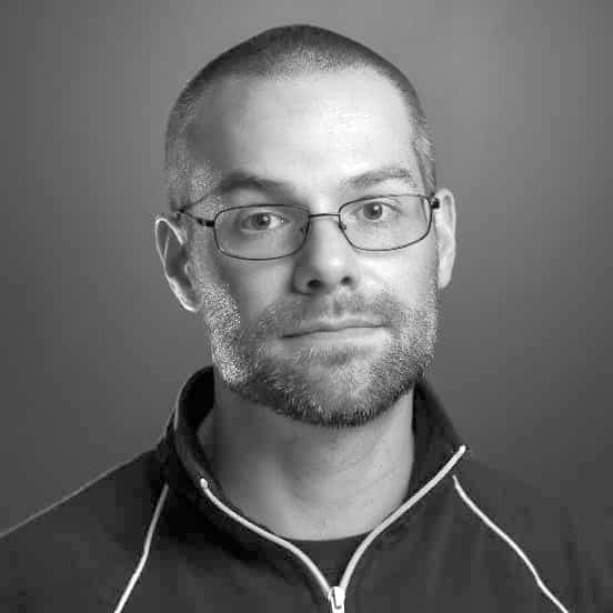 Man wearing glasses, short hair, and beard, dressed in a zipped jacket, against gray background.