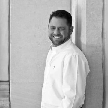 Smiling man in white shirt leaning against textured wall in black-and-white photo.