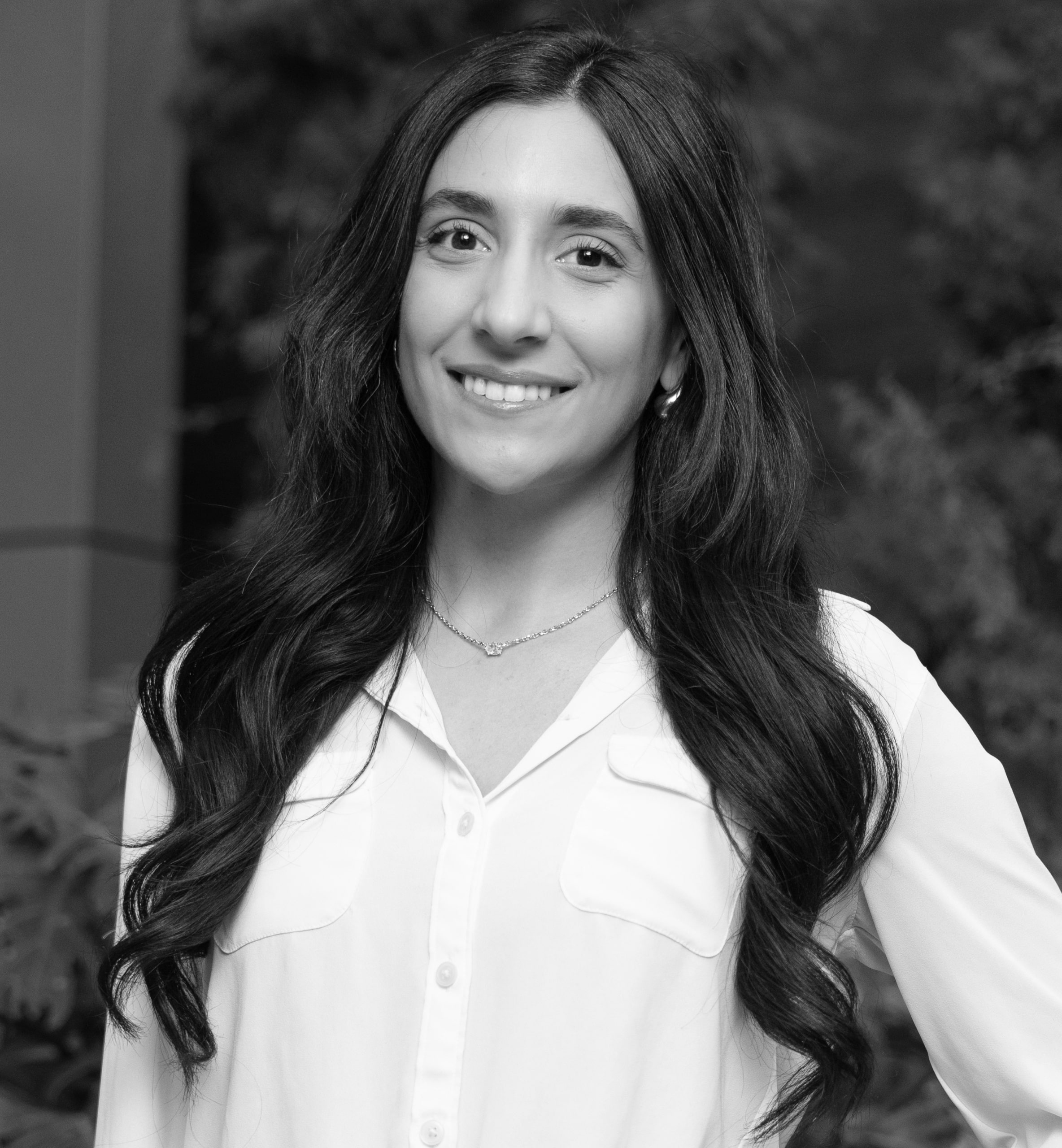 Smiling woman with long dark hair wearing a white buttoned shirt and a necklace in black and white.