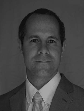 Man in a suit and tie with short hair, facing front against a plain dark background.