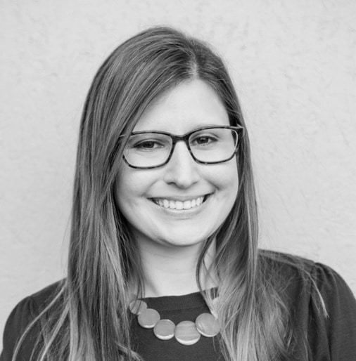 Smiling woman with long hair wearing glasses and a circular bead necklace on a plain background.