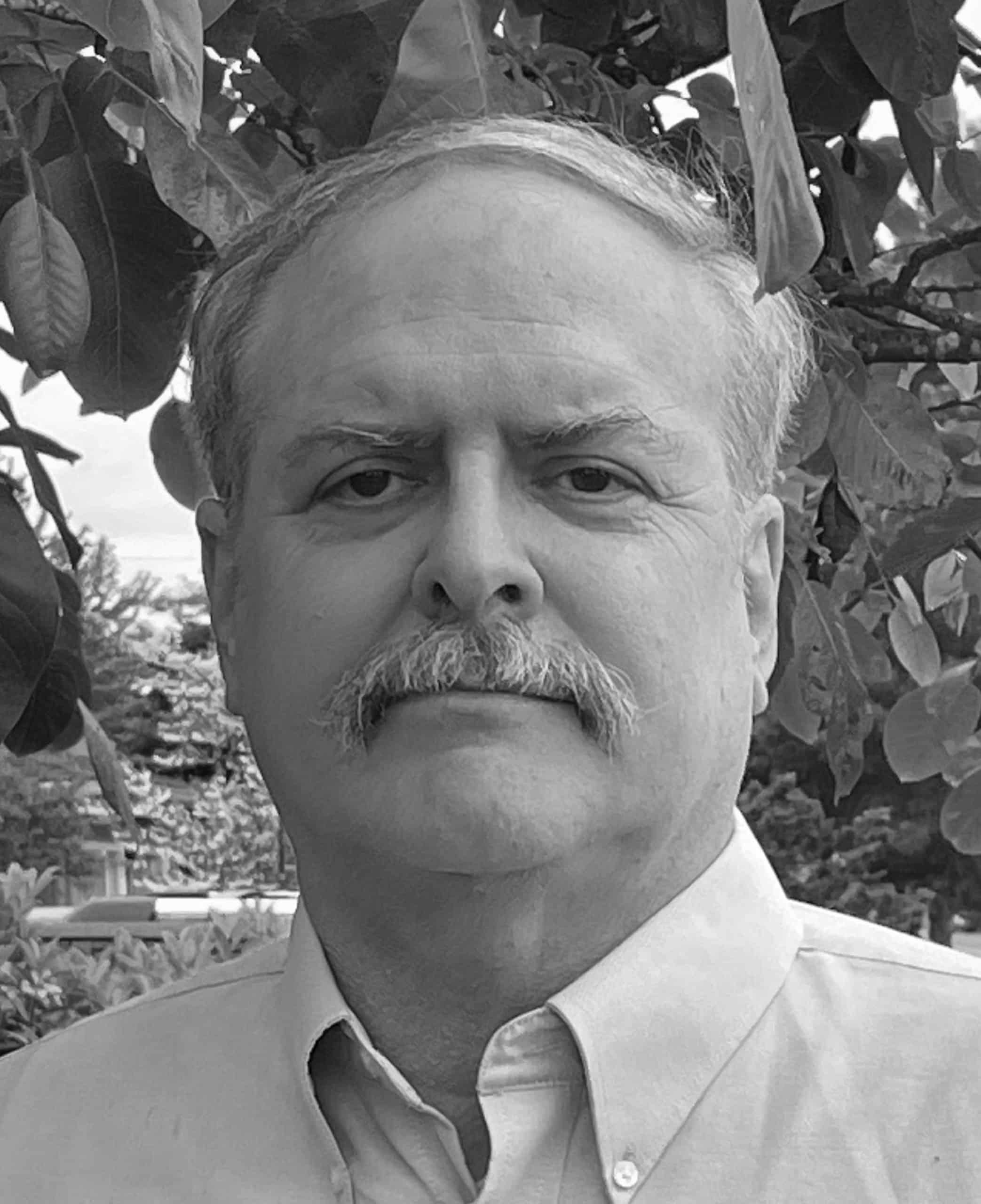 Black and white close-up portrait of a man with a mustache standing outside under tree leaves.