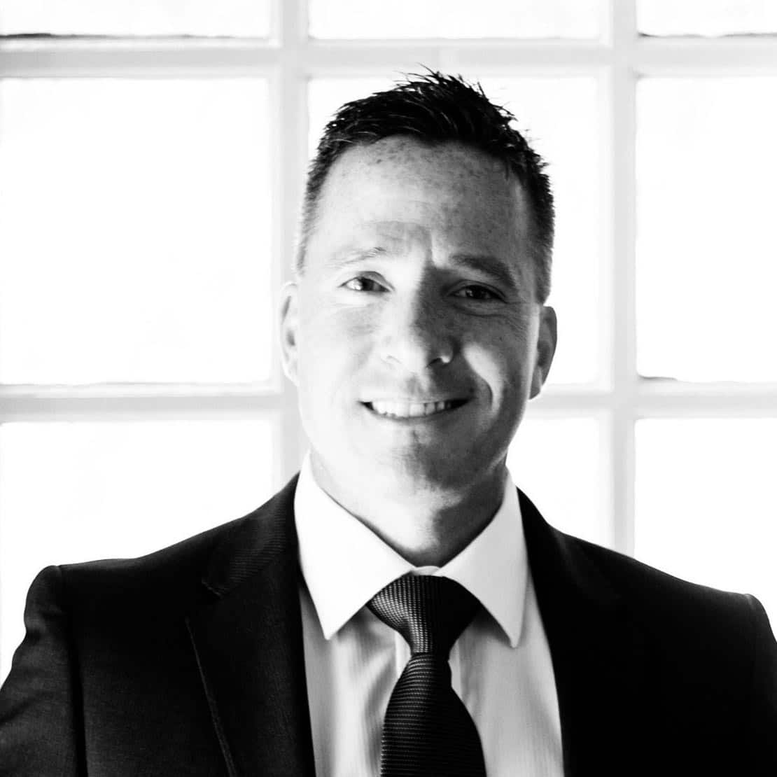 Smiling man in a suit and tie in front of a bright window with a grid pattern.