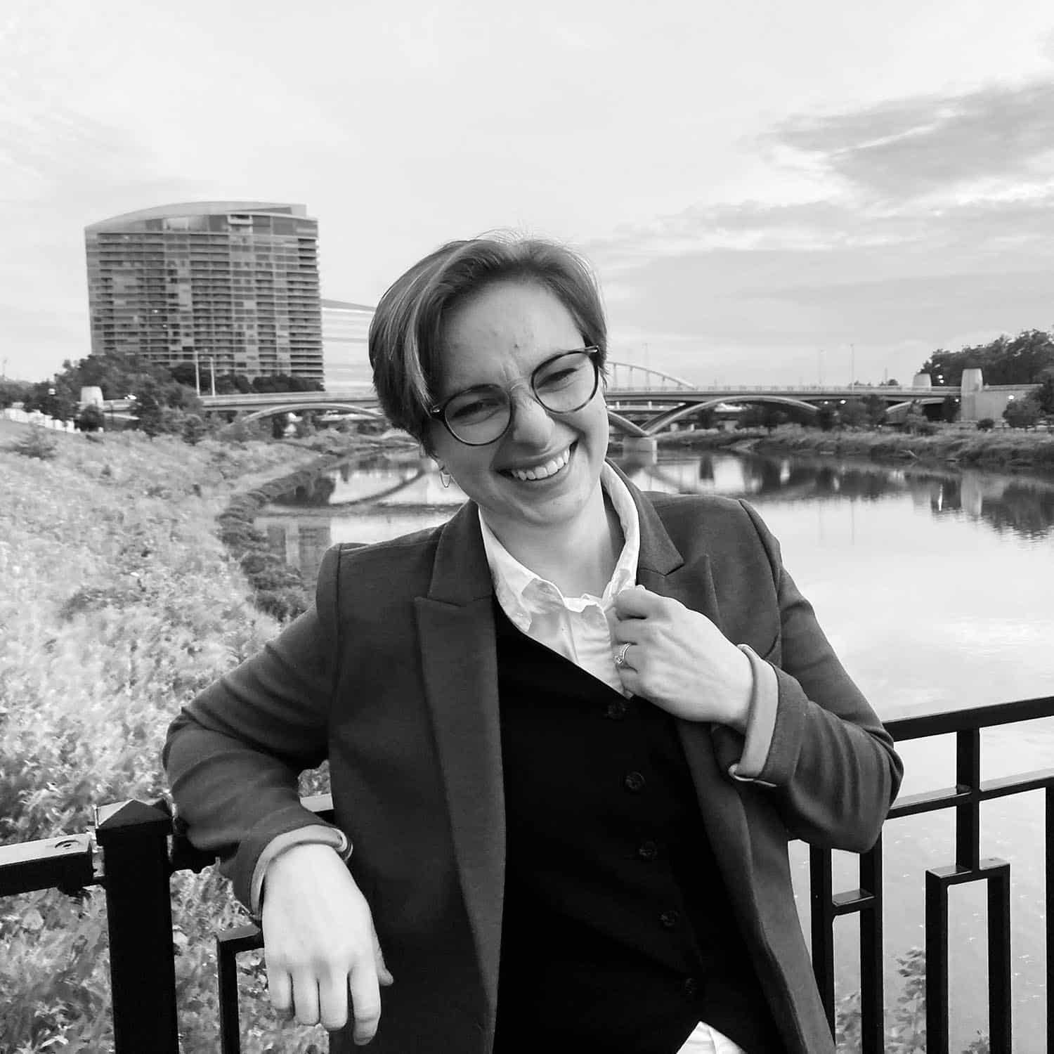 Smiling person with glasses leaning on railing by a river with a bridge and buildings in the background.