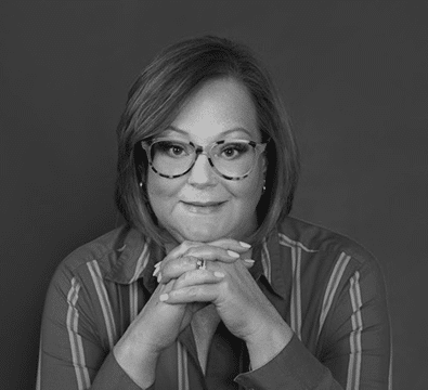 Woman with glasses and striped shirt resting chin on clasped hands, smiling slightly, on dark background.