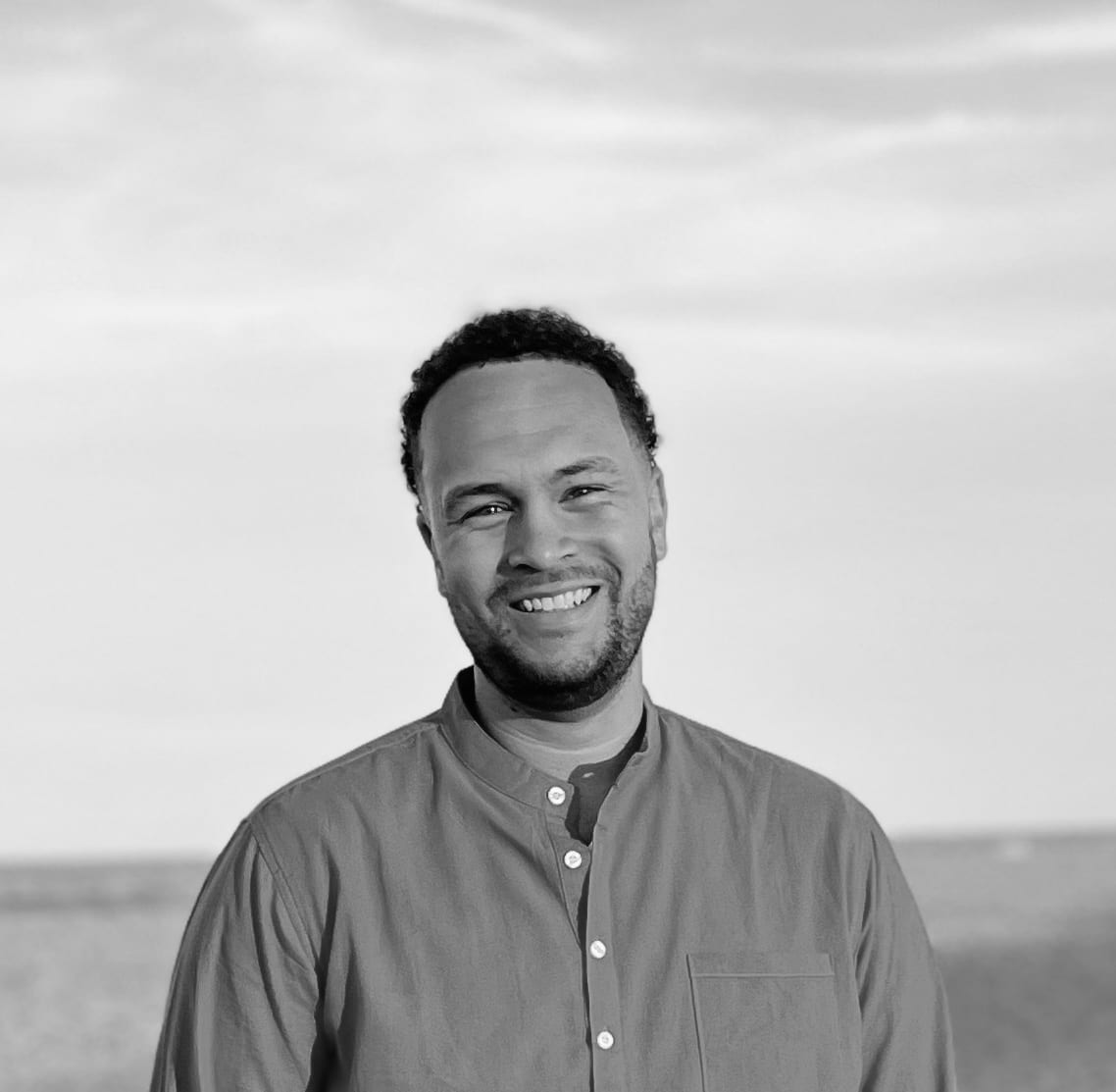 Smiling man with short curly hair and beard wearing a button-up shirt at a beach during daytime.