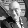 Smiling man wearing a suit and tie standing outdoors near a wooden railing.