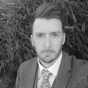 Black and white portrait of a man with short hair and beard wearing a suit and patterned tie outdoors.