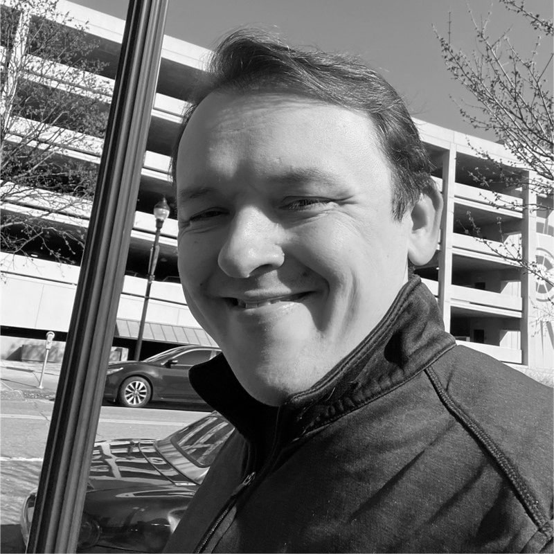 Smiling man wearing a jacket standing outdoors near a streetlamp and parked cars in daylight.
