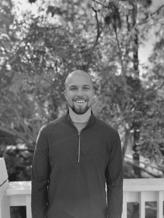 Smiling bald man with a beard, wearing a dark zip-up shirt, standing outdoors by a white railing.