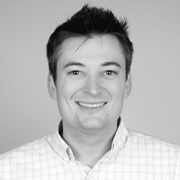 Smiling young man with short dark hair wearing a buttoned-up checkered shirt on a plain background.