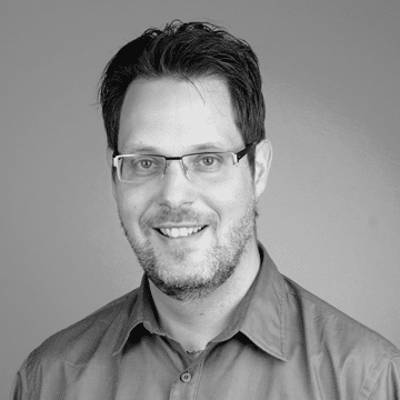 Headshot of a man with glasses, short dark hair, and a beard, smiling in front of a plain background.