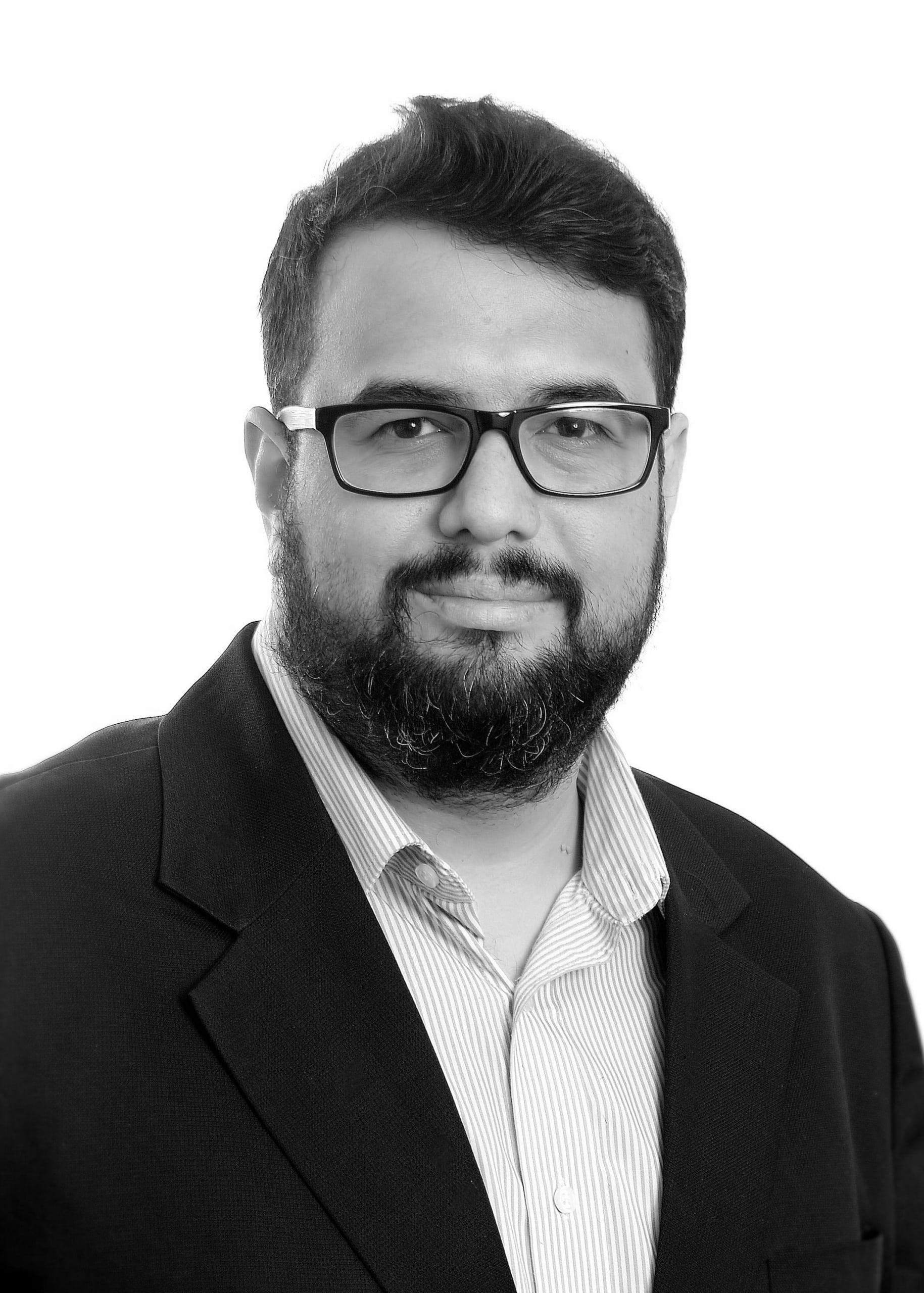 Man with dark hair, beard, and glasses wearing a striped shirt and dark blazer on white background.