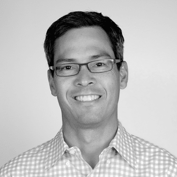 Smiling man wearing glasses and a checkered button-up shirt against a plain background.