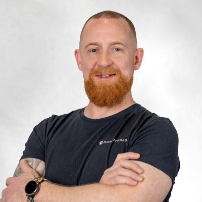 Man with red beard, crossed arms, wearing black Domtools shirt and watch, smiling at camera.