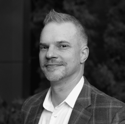 Smiling man with short spiked hair wearing a plaid blazer and white shirt in black and white.