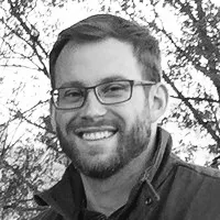 Smiling man with glasses and beard wearing a jacket outdoors with tree branches in background.