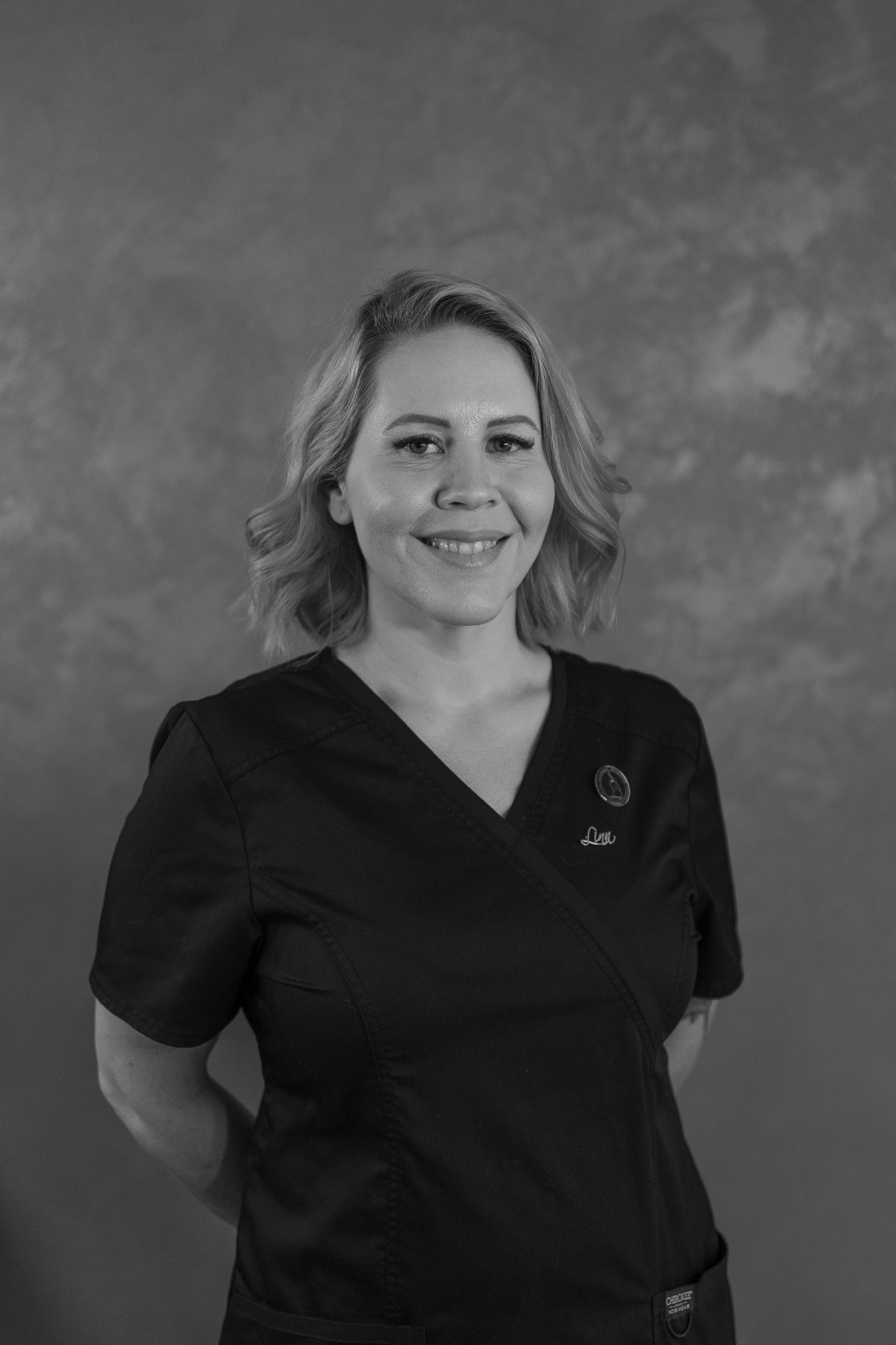 Smiling woman with shoulder-length hair in a black scrub top against plain background.