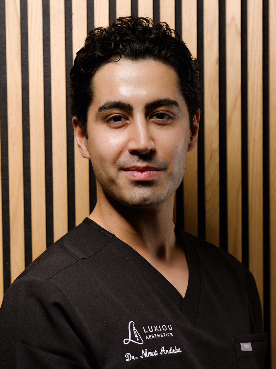 Portrait of a man with curly hair wearing black scrubs embroidered with Luxiou Aesthetics and Dr. Nimat Andisha.