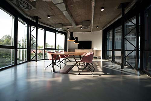 Modern conference room with large windows, a wooden table, and pink chairs under hanging lights.