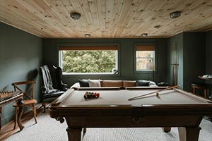 Cozy game room with a wooden pool table, cue sticks, a cushioned bench under two windows, and wooden ceiling and furniture.