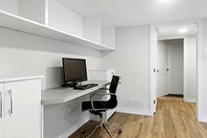 Minimalist home office setup with a desk, computer monitor, black swivel chair, white cabinets, and light wooden floor.