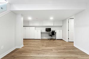 Minimalist basement room with light wood flooring, white walls, built-in desk with computer, and overhead cabinets.