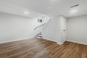 Empty white basement room with wooden floor, staircase with railing, and closed door.