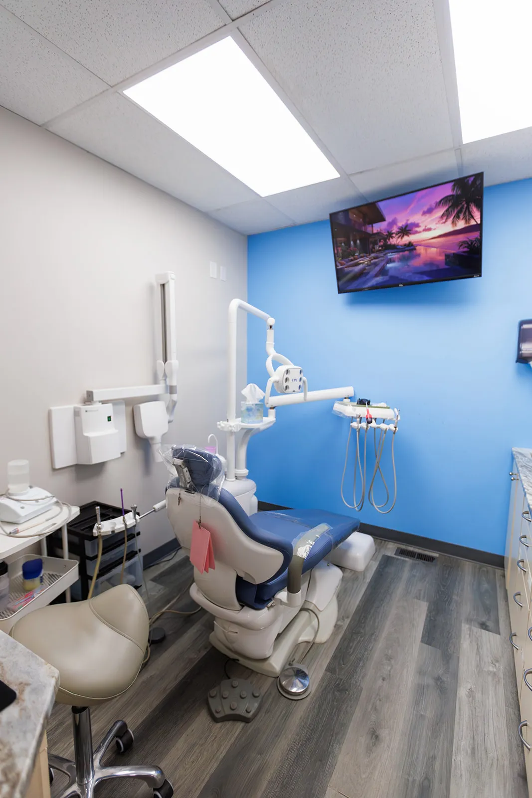 Marvel dental examination room with blue dental chair, medical equipment, and a wall-mounted TV displaying a tropical sunset scene.