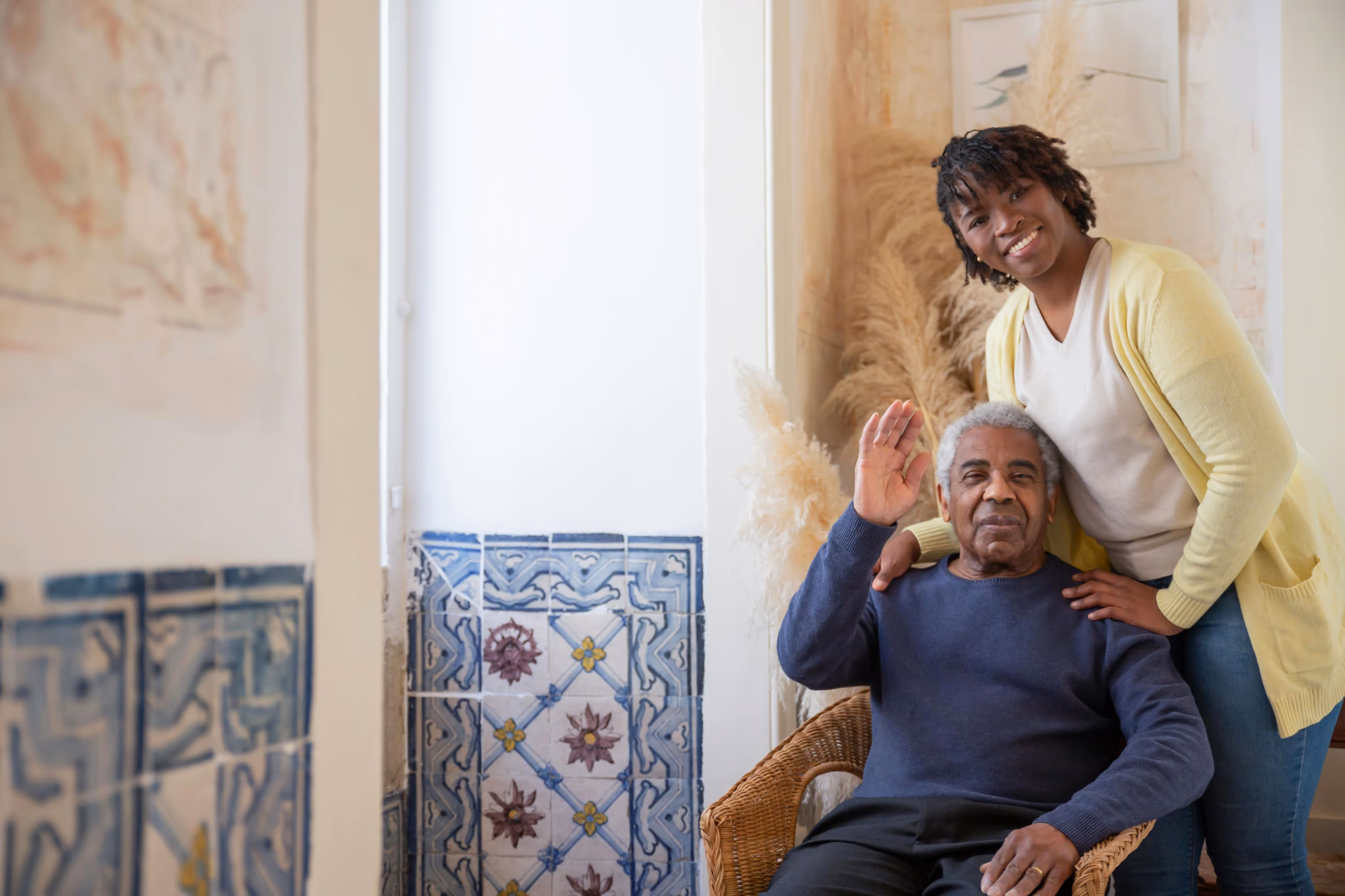 Smiling young woman standing behind an elderly man in a wicker chair who is waving, in a warmly decorated room with patterned tiles.