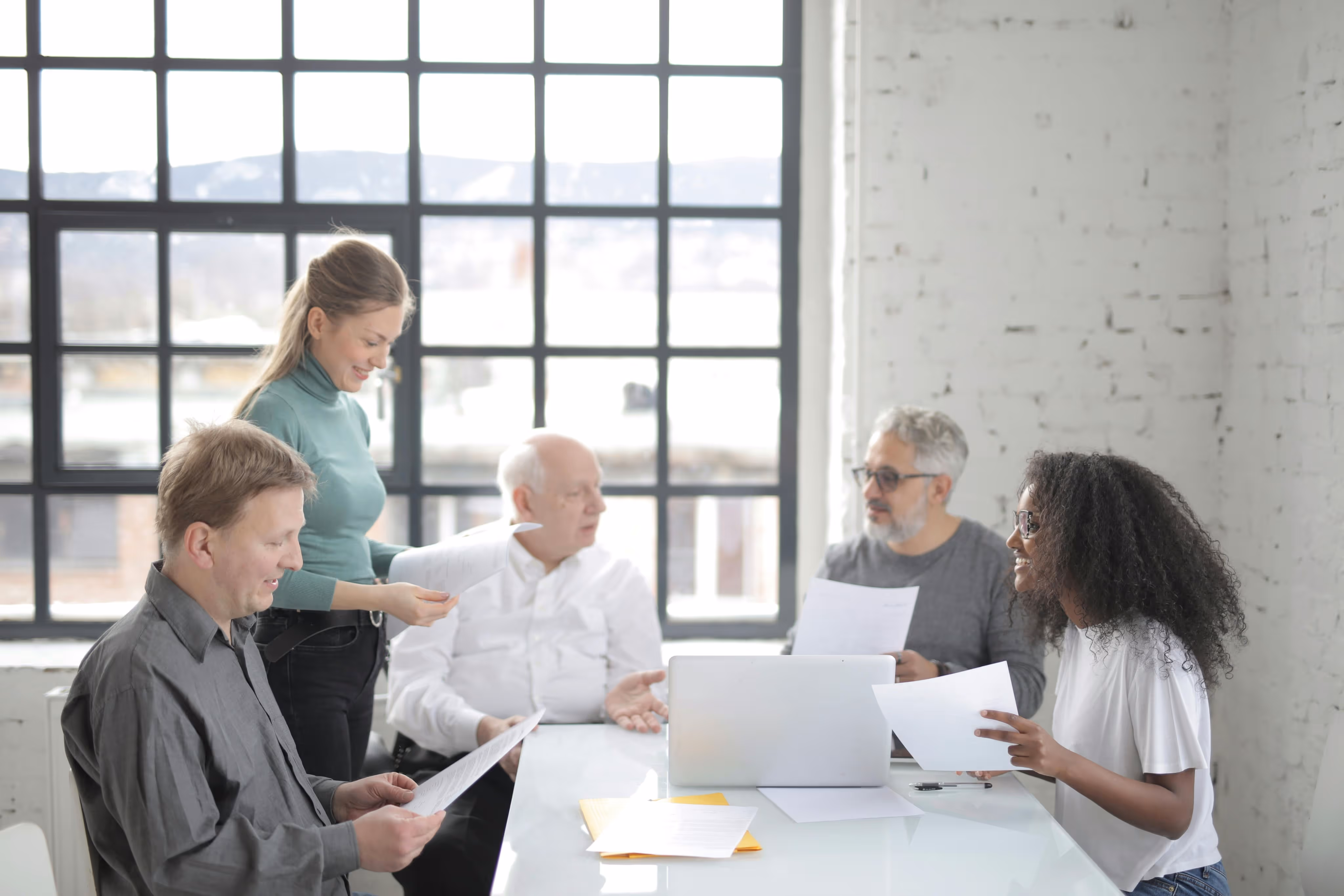 Five diverse colleagues reviewing documents and discussing around a conference table with a laptop in a bright office.