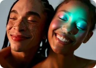 Close-up of two smiling young women with eyes closed, one with freckles and the other with radiant skin under colorful lighting.
