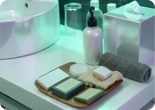 Bathroom counter with a sink, tissue box, pump bottle, rolled gray towel, and wooden tray holding soap bars and cotton pads.