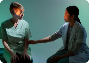 Two healthcare professionals sitting and having a supportive conversation, one gently holding the other's arm.
