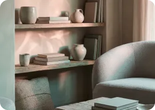 Cozy reading nook with a curved light gray armchair, wooden shelves holding books and ceramic vases, and soft natural light.