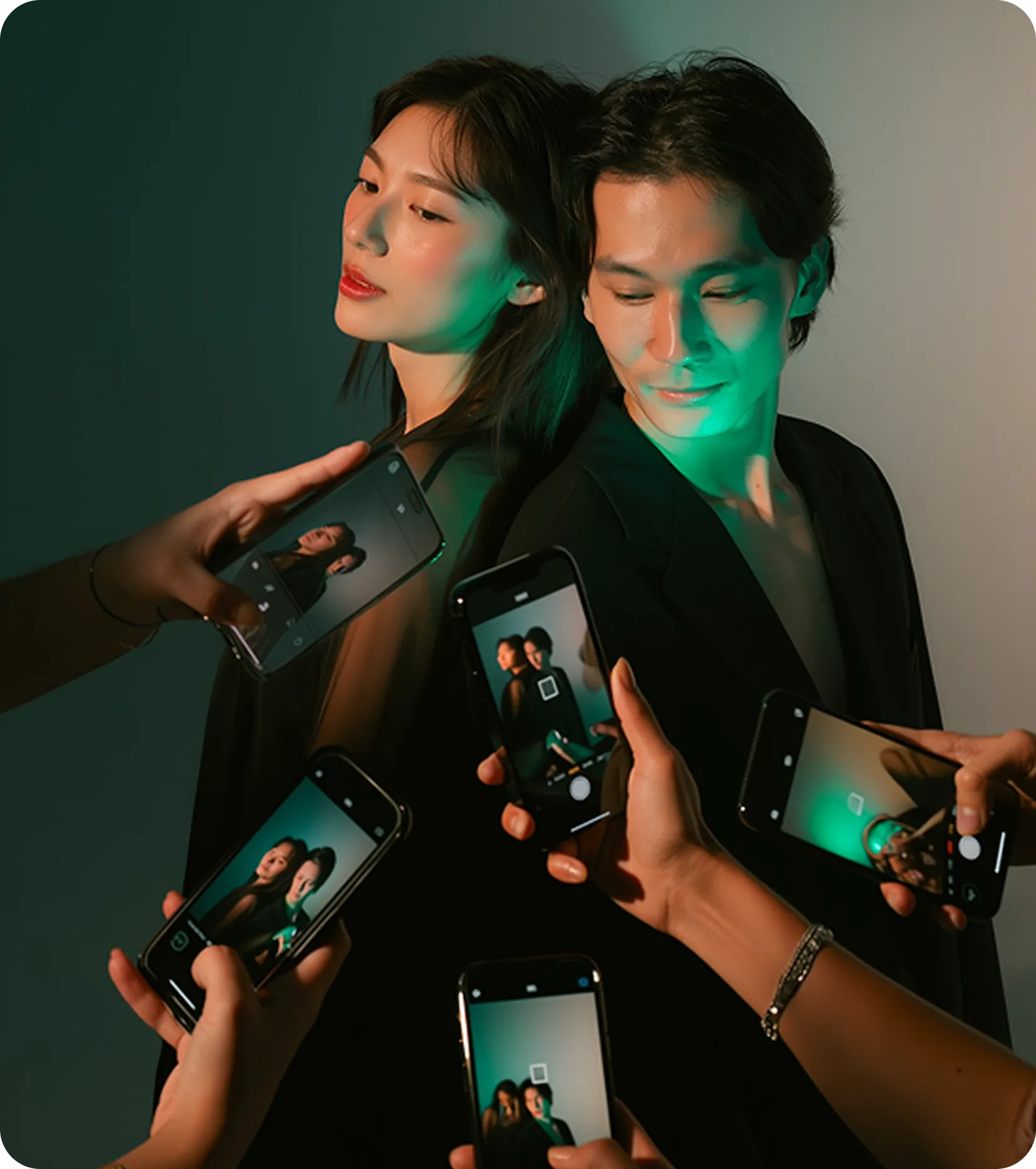 Two people posing back-to-back with multiple phones photographing them in a dimly lit setting with green lighting.