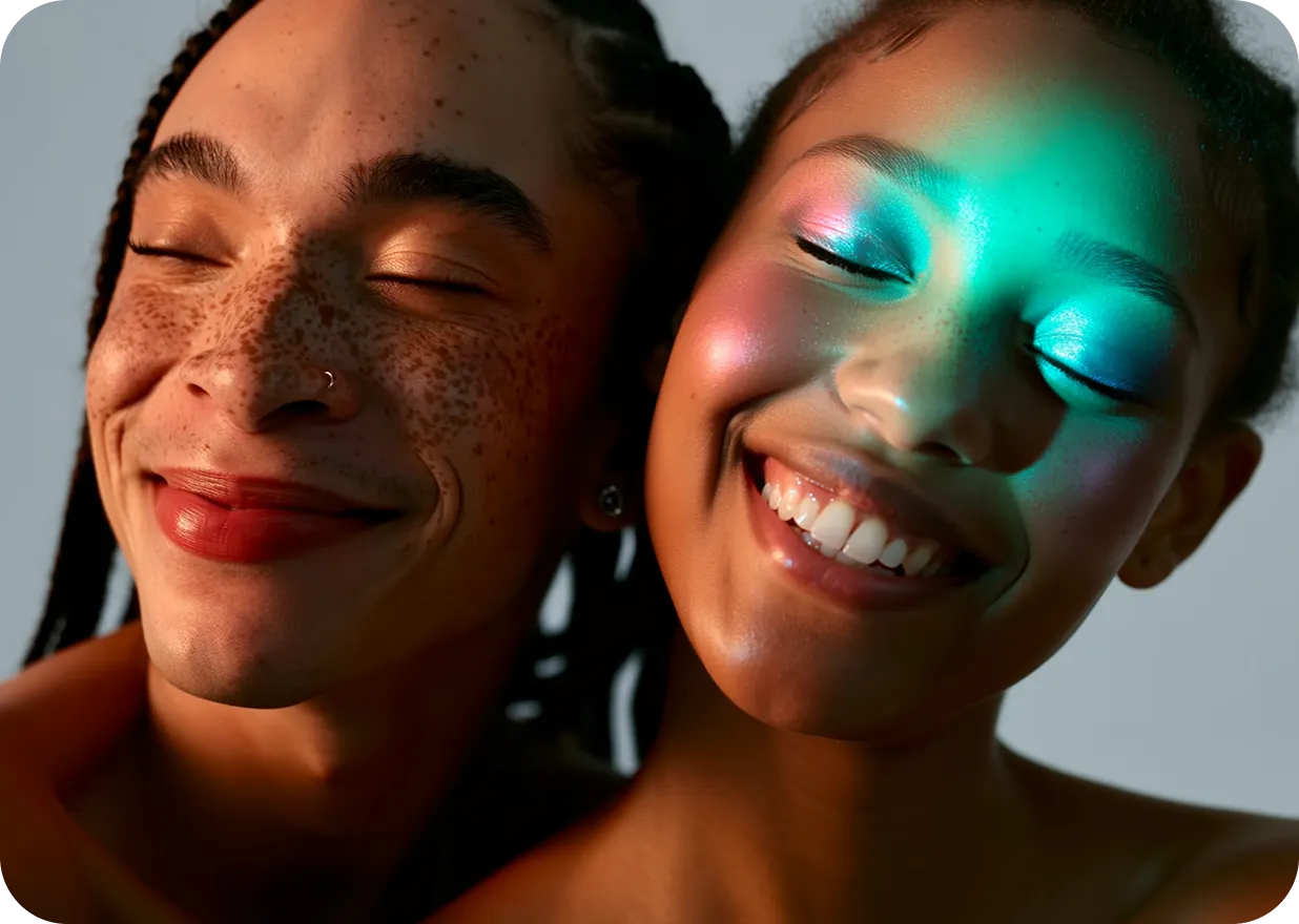 Close-up of two people with eyes closed smiling, one with freckles and nose ring, the other with colorful light on their face.