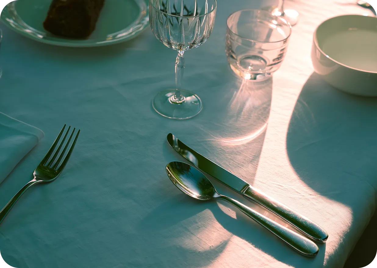 Table setting with a fork, knife, spoon, crystal wine glass, water glass, white bowl, and a white plate with a piece of bread on a white tablecloth.