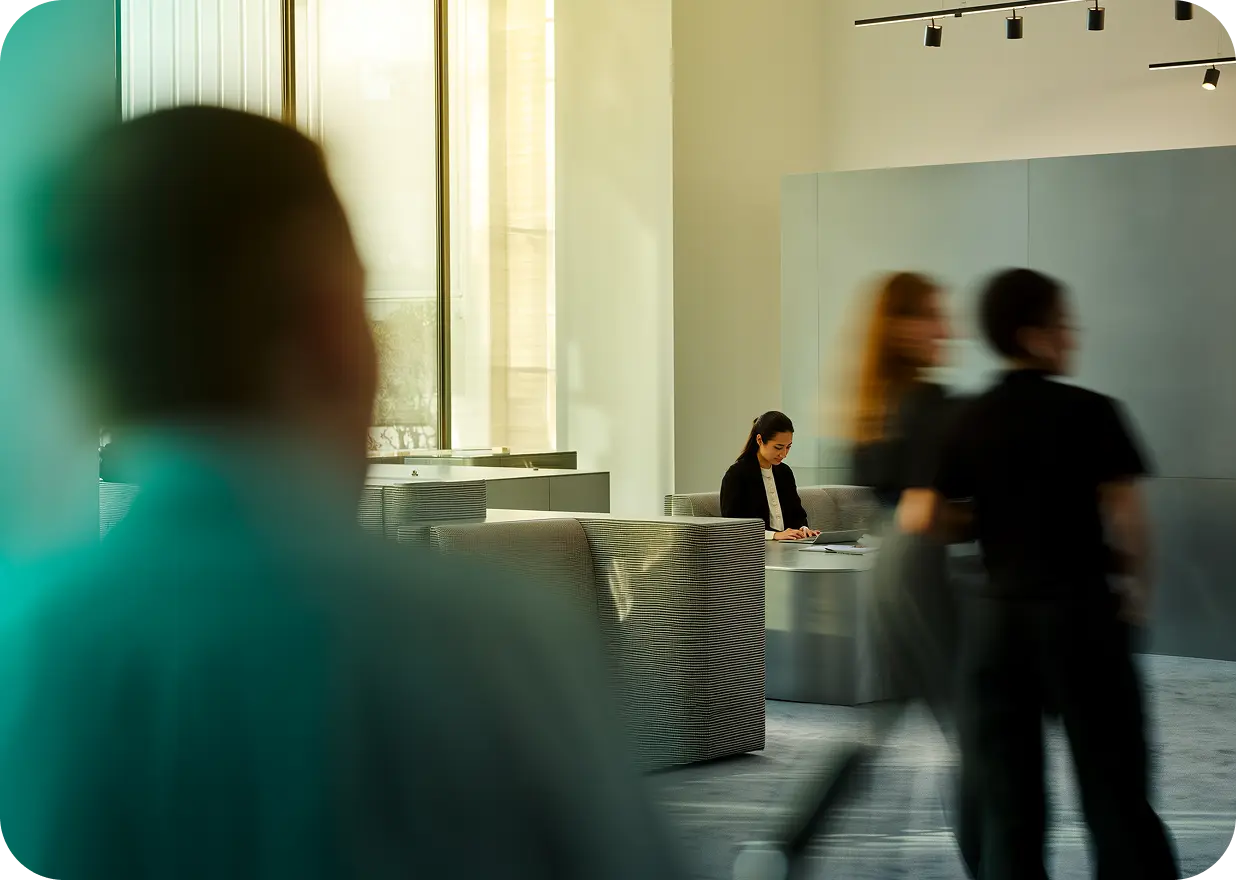 Woman sitting alone on a modern sofa in a bright office lobby, working on a laptop while blurred figures walk past.