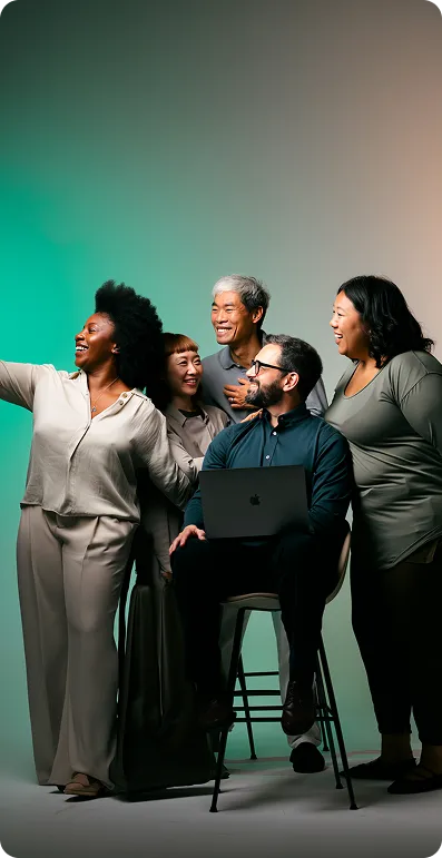 Diverse group of five colleagues smiling and gathering for a selfie, with one person seated holding a laptop.