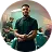 Confident man with folded arms standing in a green-lit environment with blurred people in the background.