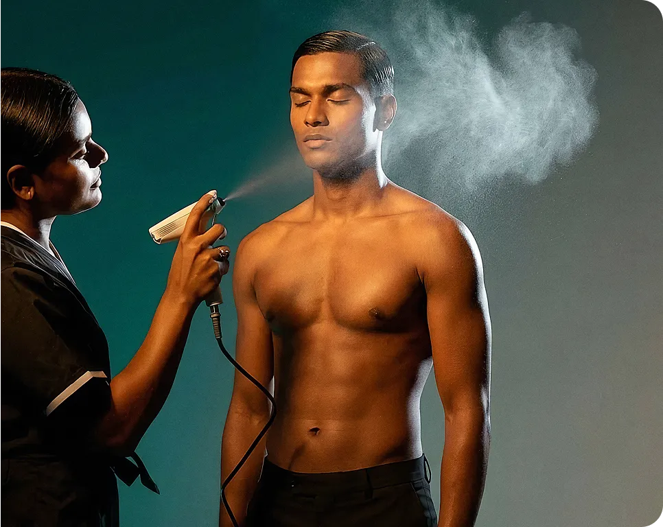 A woman sprays a mist on the face of a shirtless man with eyes closed, against a dark background.