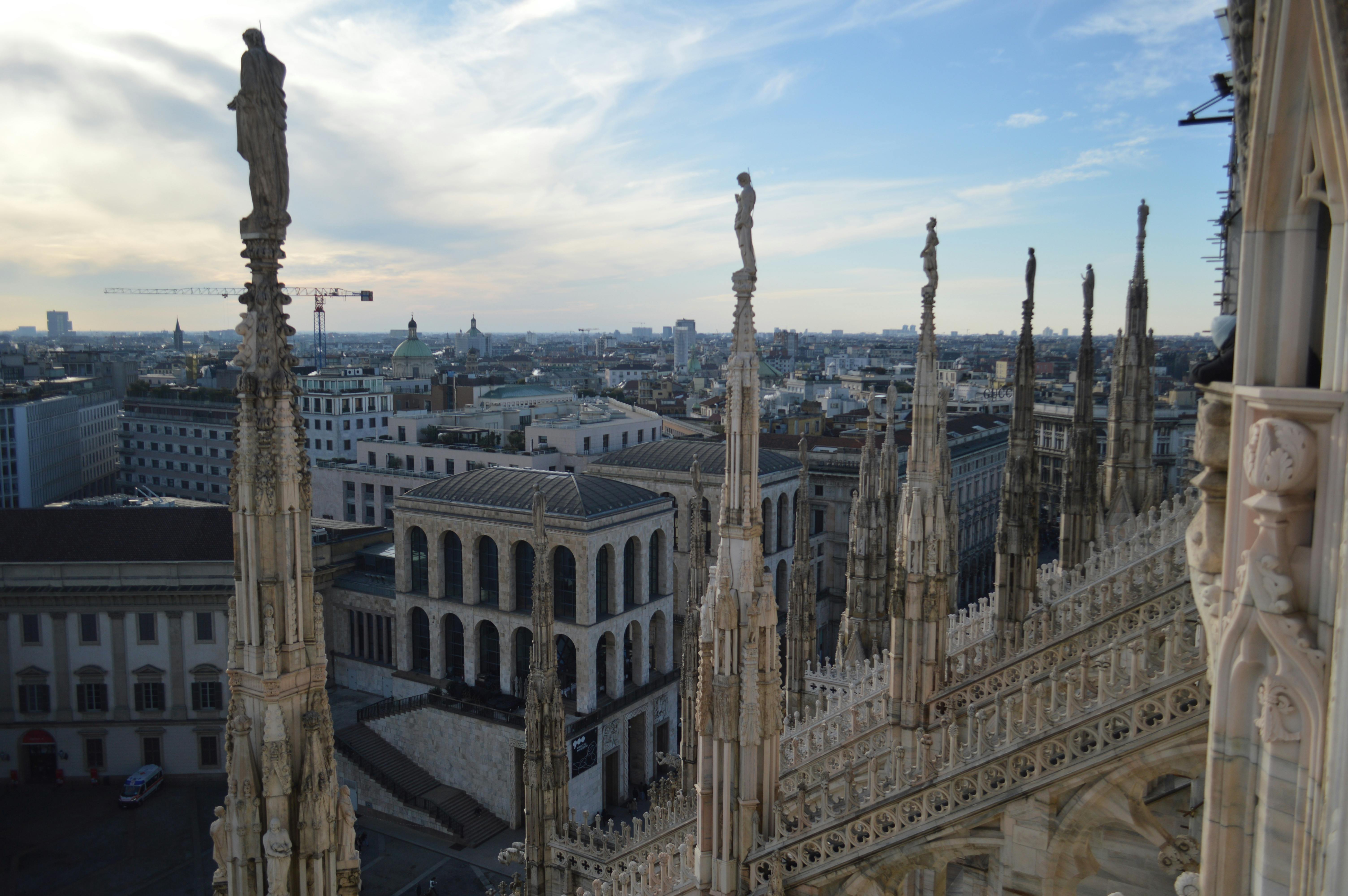 View from Milan Cathedral rooftop showing Gothic spires and statues with city buildings and sky in the background.