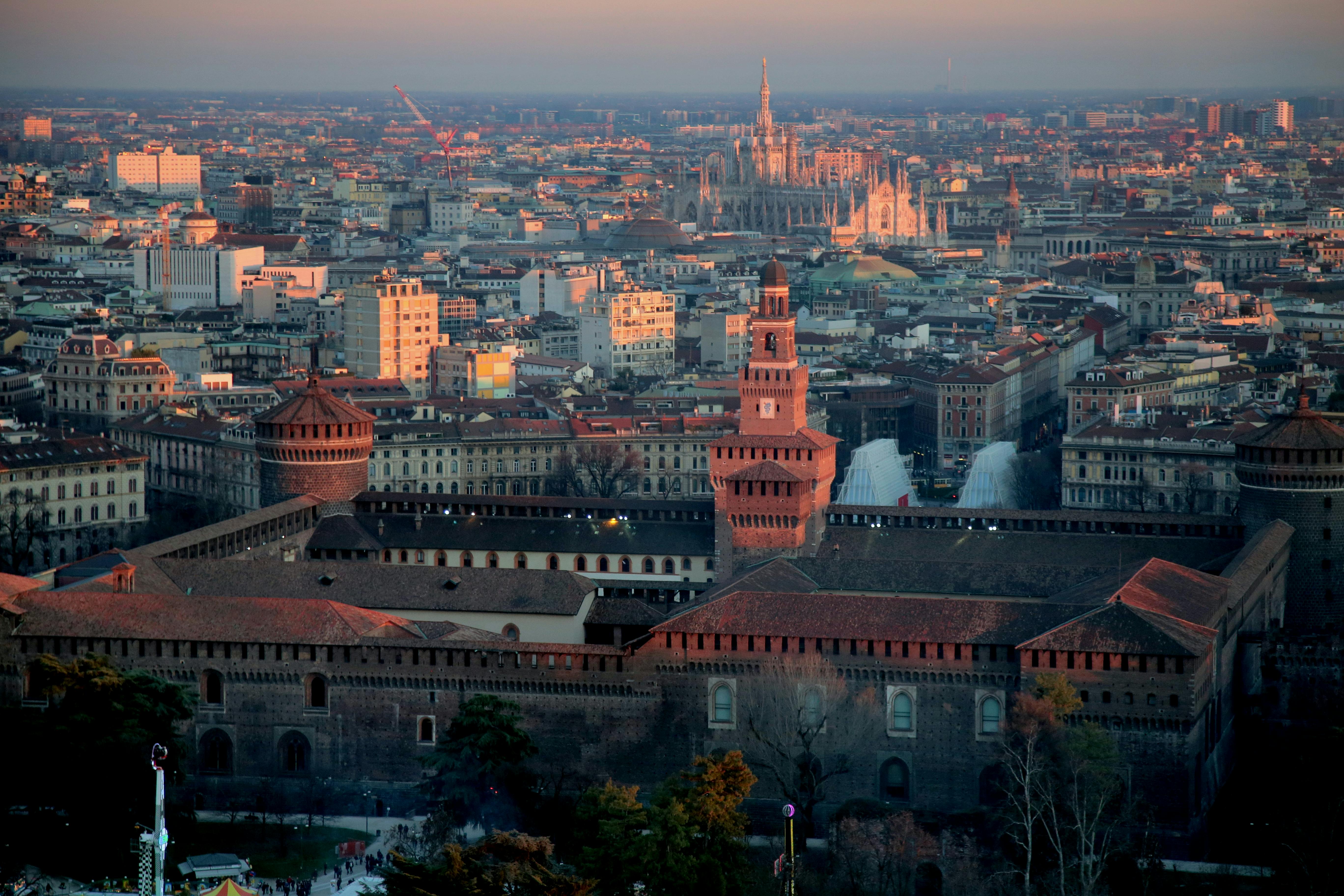 Aerial view of Castello Sforzesco with Milan cityscape and the illuminated Milan Cathedral in the background at sunset.