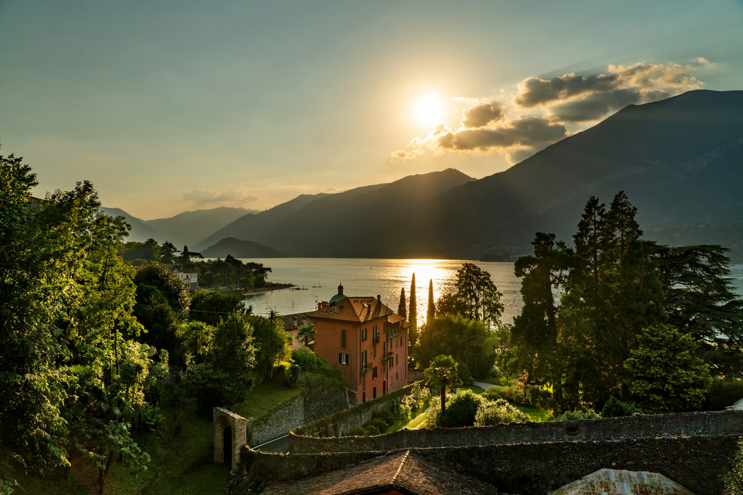 Sunset over Lake Como with mountains, a large orange building, and lush green trees.