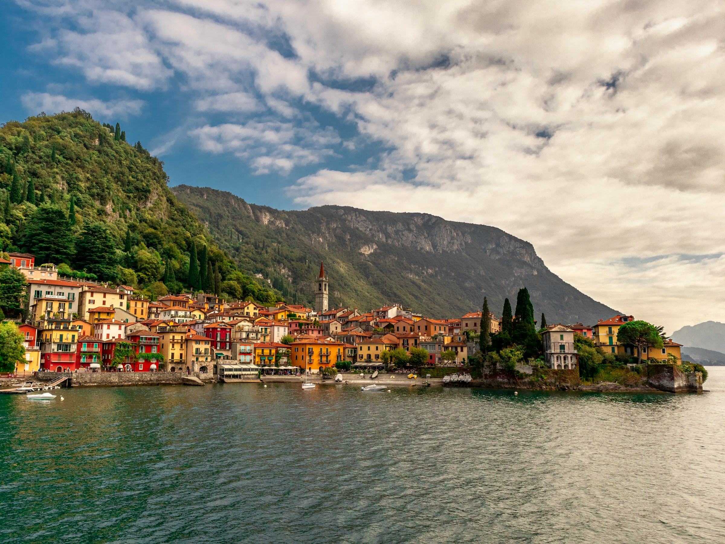 Colorful lakeside village with church tower nestled at the foot of green mountains under a partly cloudy sky.