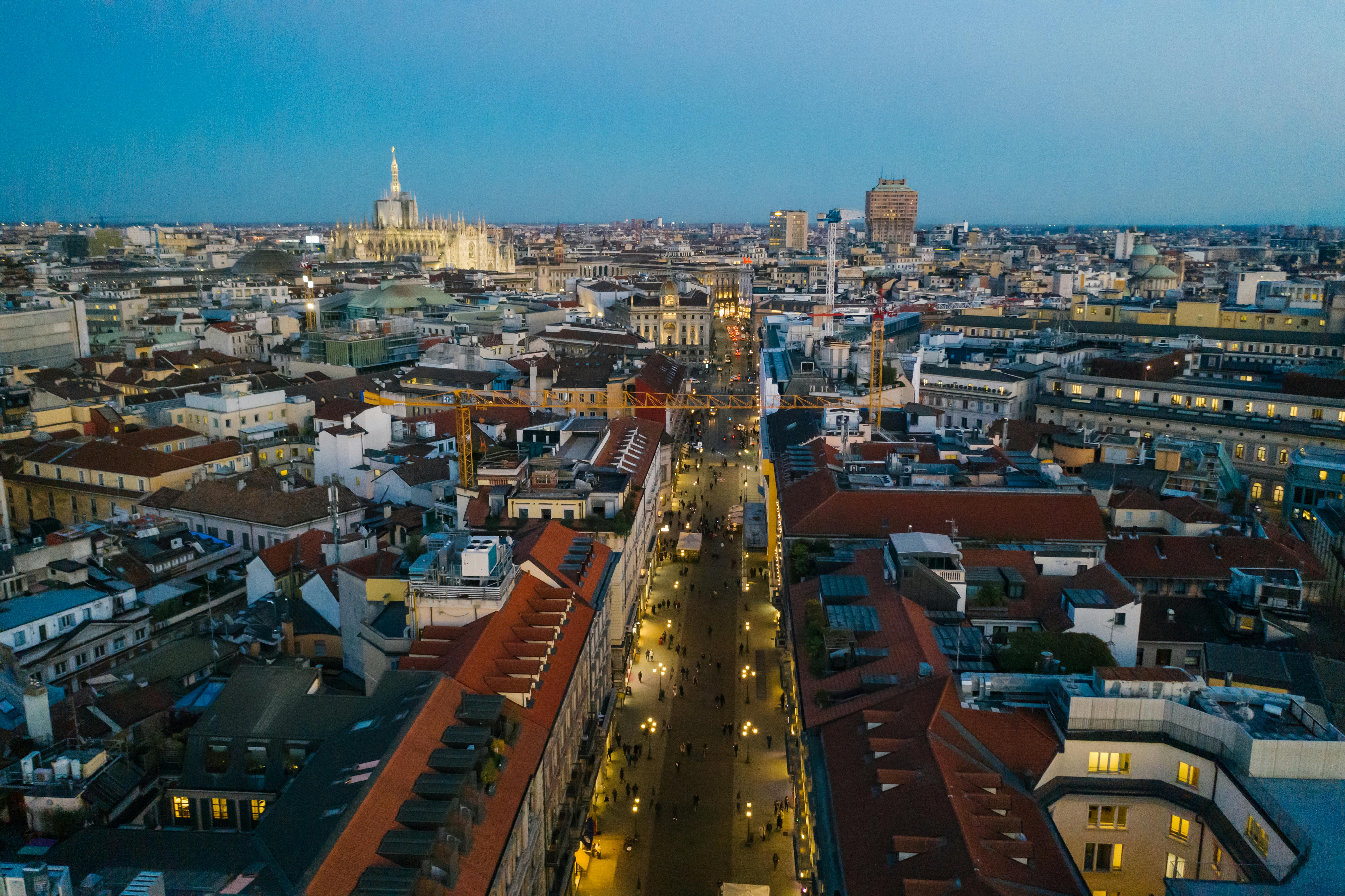 Aerial view of Milan city at dusk with the illuminated Milan Cathedral and a busy pedestrian street lined with buildings.